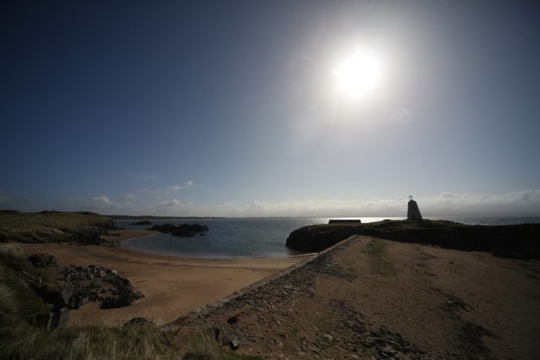 Coastal landscape with sun shining over a beach, rocky terrain in the foreground, and a small lighthouse in the distance.