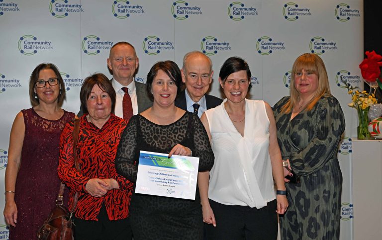 Group of seven people posing with a certificate at an awards ceremony, standing in front of a banner with logos.