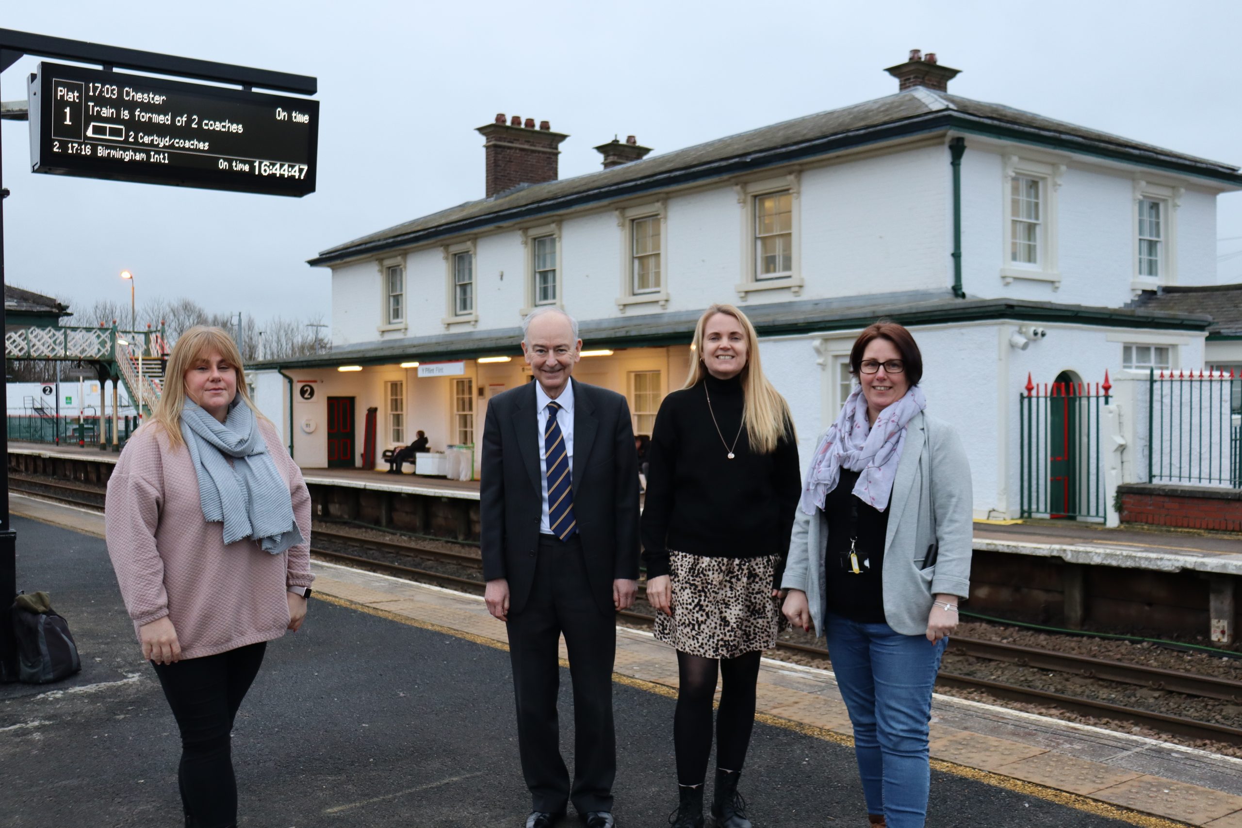 Four people standing on a train station platform, with a train schedule display and the station building in the background.