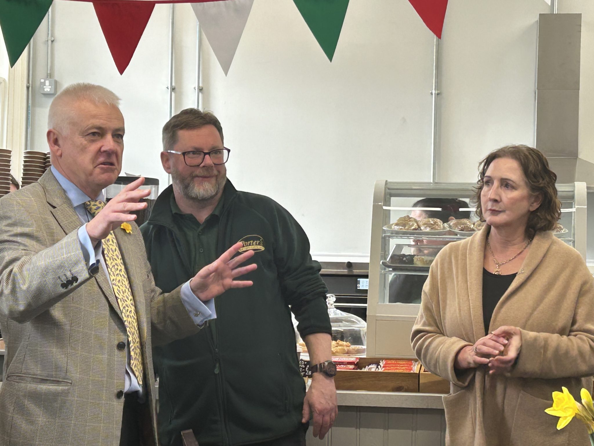 Three people standing in a cafe with one man speaking and gesturing, another listening attentively, and a woman looking on calmly, with red, white and green bunting in the background.