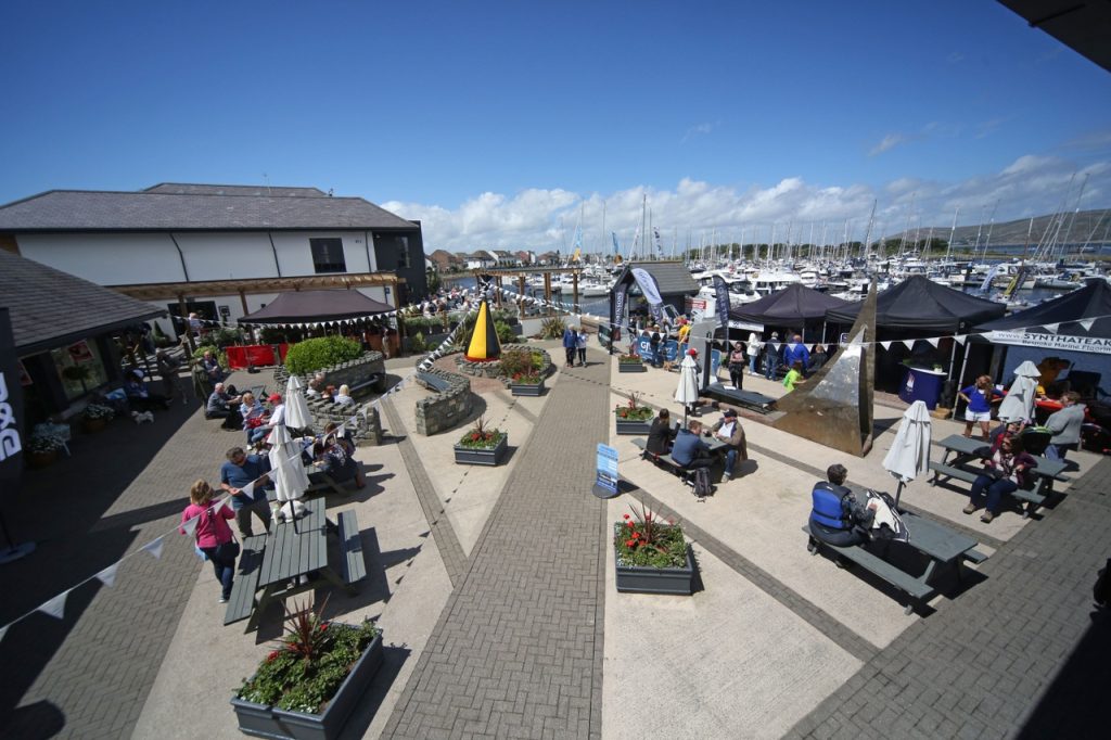 A bustling marina with people dining at outdoor tables, various boats docked nearby, and tents set up along the walkway under a clear blue sky.