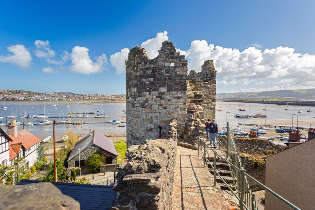 Ancient stone castle ruins overlooking a harbor with boats, under a clear blue sky with scattered clouds. Visitors explore the historic site on a sunny day.