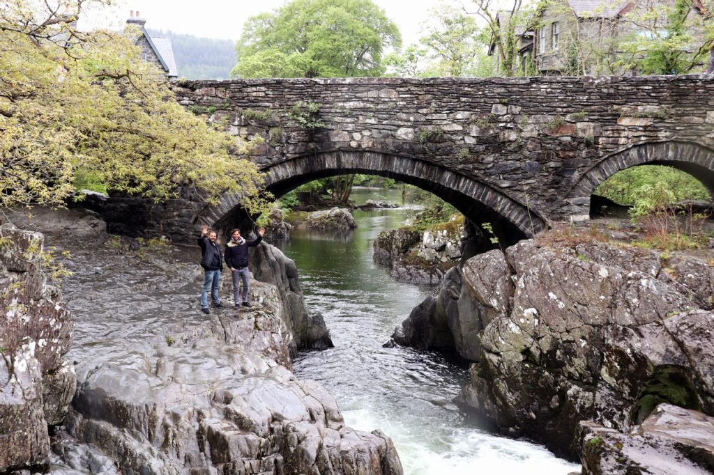 Two individuals waving by a rocky river under an old stone arch bridge with trees and greenery in the background.