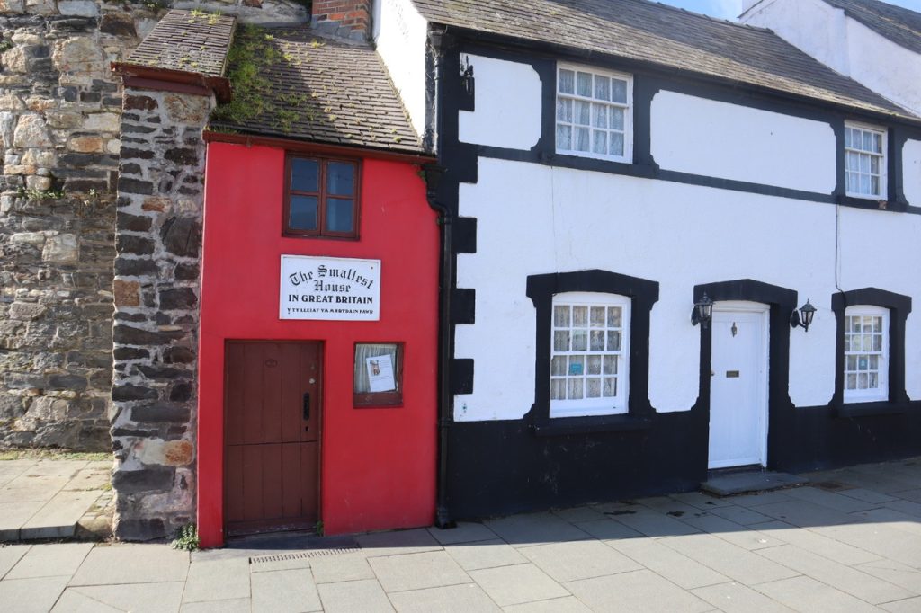 A narrow, red two-story building with a distinctive sign proclaiming it 'The Smallest House in Great Britain,' nestled between larger buildings.