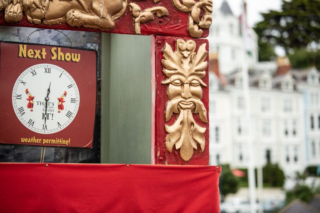 A clock with the text "Next Show" above it and "weather permitting!" below, mounted on a wall with decorative carvings, including a gold-colored mask motif, beside a red curtain.