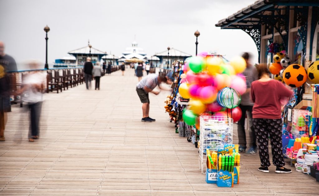A person browsing items at a stall on a wooden pier with colorful balloons and toys for sale in the foreground. The pier extends into the background with other visitors walking along its length.