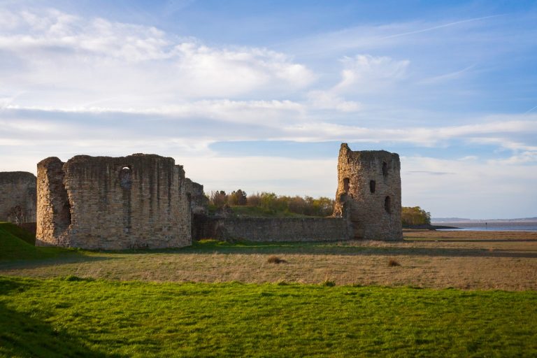 Ruins of an old stone castle with towers against a clear sky, surrounded by an open grassy field.