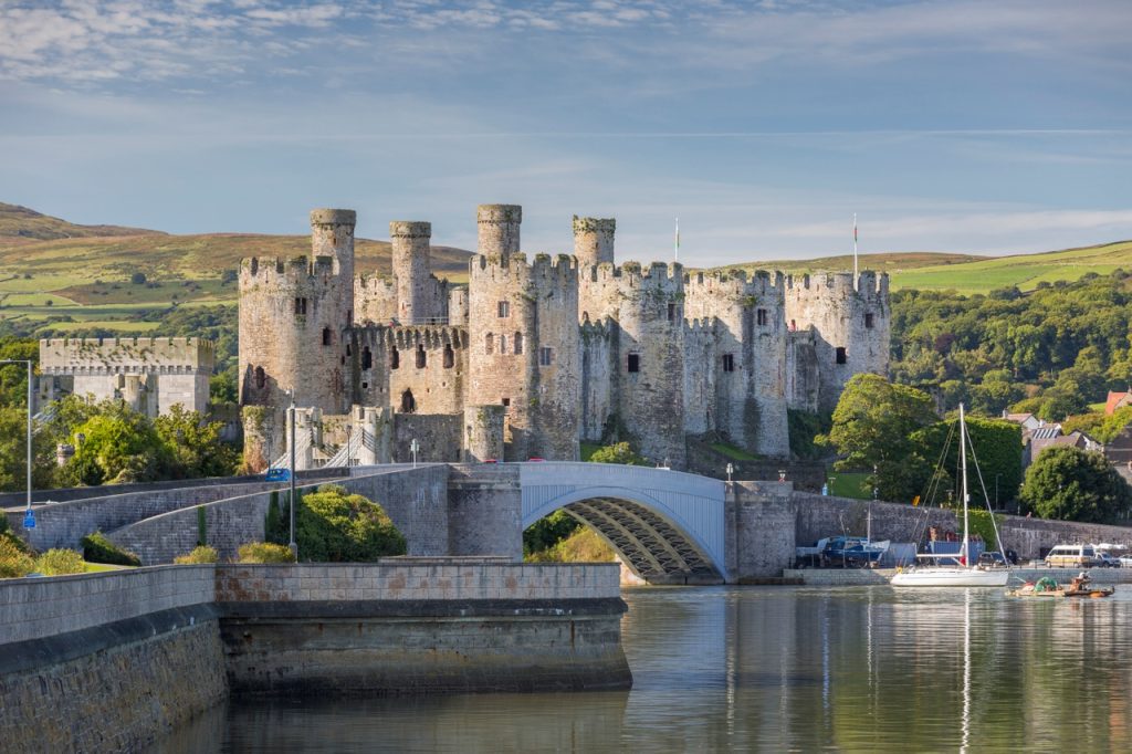 Historic medieval castle with multiple towers by a river with an arched stone bridge in the foreground.