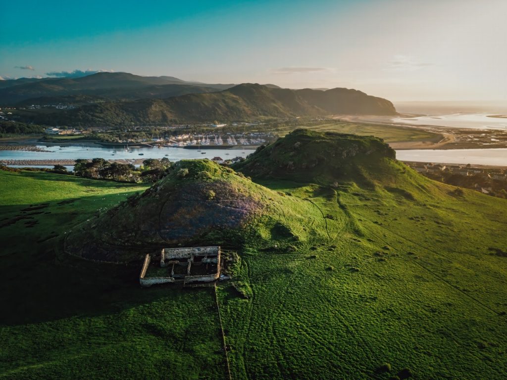Aerial view of a lush green hill with ruins overlooking a coastal town during sunset.