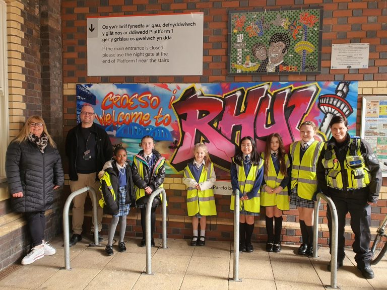 Group of children wearing high-visibility vests and three adults standing in front of a board that says "Croeso i / Welcome to RHYL" in graffiti writing at a railway station.