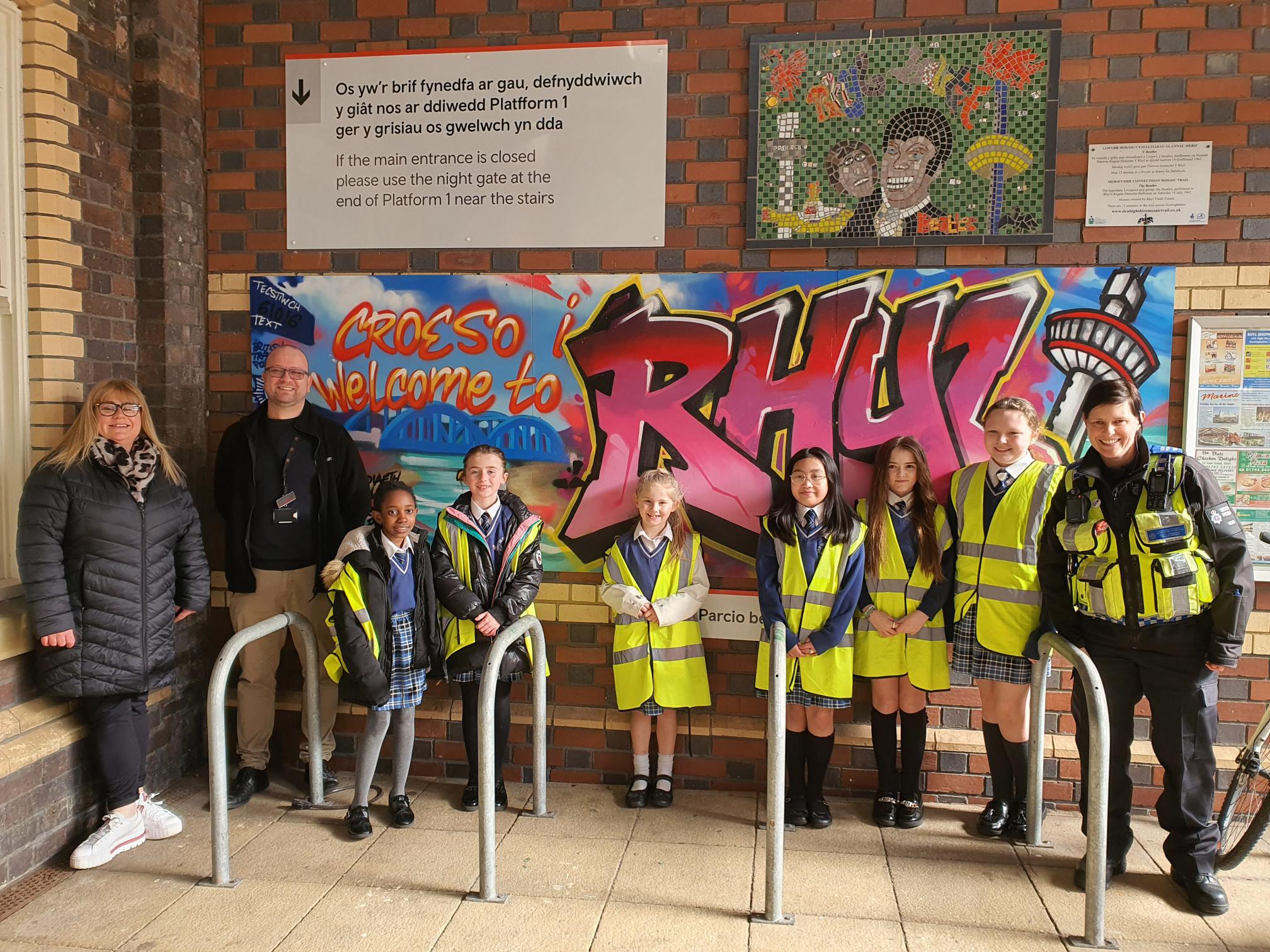 Group of children wearing high-visibility vests and three adults standing in front of a board that says "Croeso i / Welcome to RHYL"  in graffiti writing at a railway station.