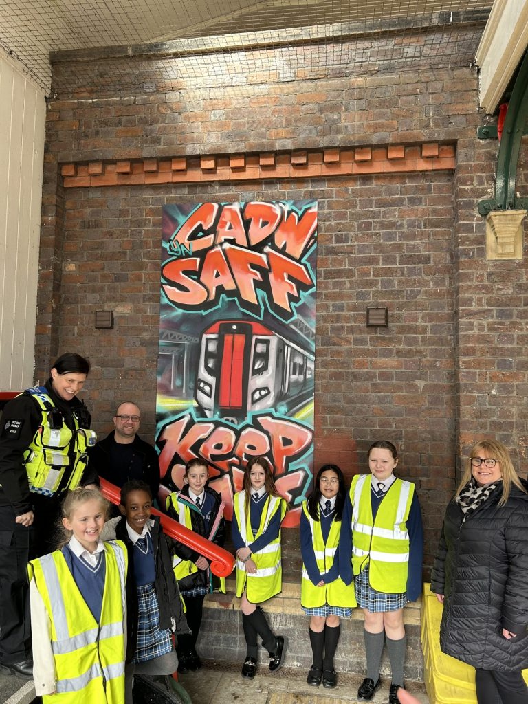 Group of children wearing high-visibility vests and three adults standing in front of a board that says "CADW SAFF KEEP SAFE" with a train in the middle in graffiti writing at a railway station.